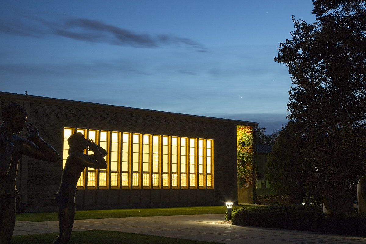 Evening view of Cranbrook Academy of Art Library. A long, horizontal brick building, mostly in shadow, with a row of vertical windows across most of the facade. Golden light emanates from the interior. Some dark tree and foliage shapes are off to the right